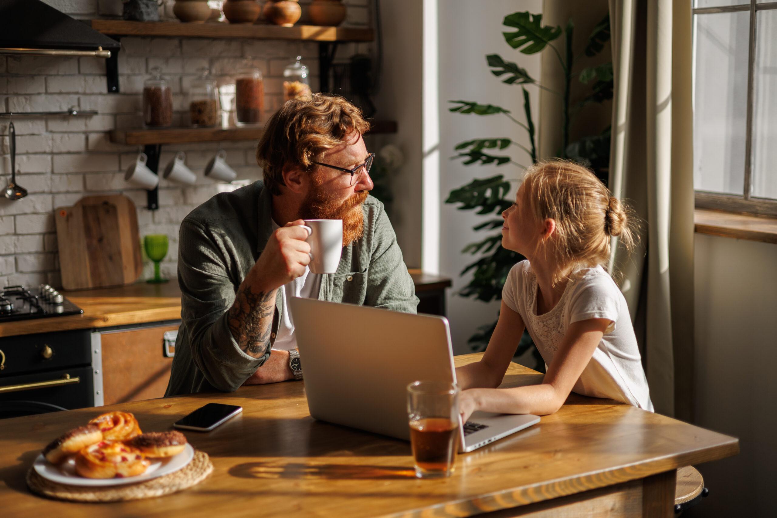 Father holding coffee cup and talking to smiling daughter using laptop during breakfast in kitchen