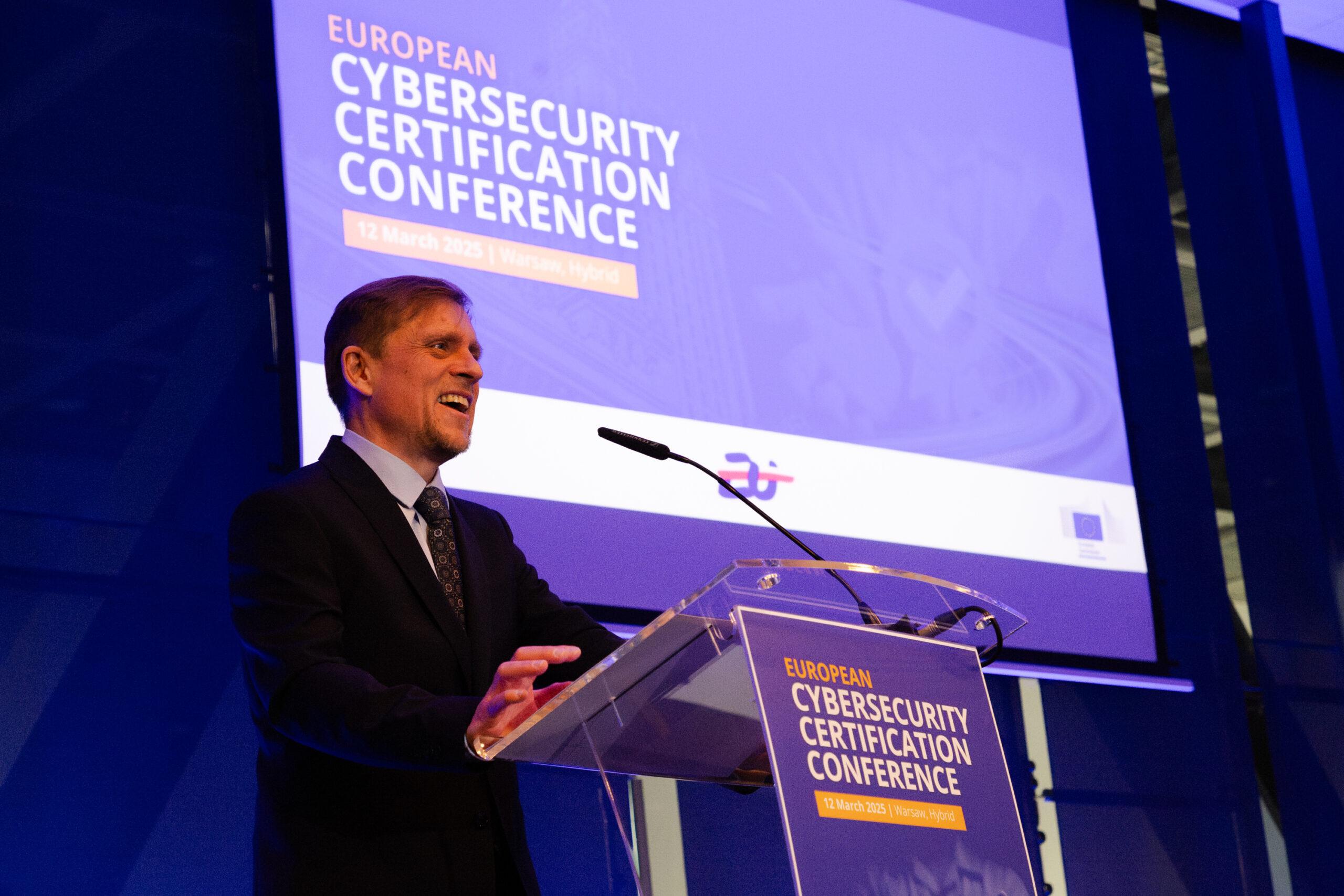 Na zdjęciu widać mężczyznę w garniturze przemawiającego na konferencji. Stoi za przezroczystą mównicą z logotypem i napisem "EUROPEAN CYBERSECURITY CERTIFICATION CONFERENCE", z datą 12 March 2025 | Warsaw, Hybrid. W tle widoczny jest duży ekran z tym samym tytułem wydarzenia oraz logotypami instytucji, w tym Unii Europejskiej. Scena jest profesjonalnie oświetlona, a prelegent wygląda na uśmiechniętego i zaangażowanego w swoją wypowiedź.