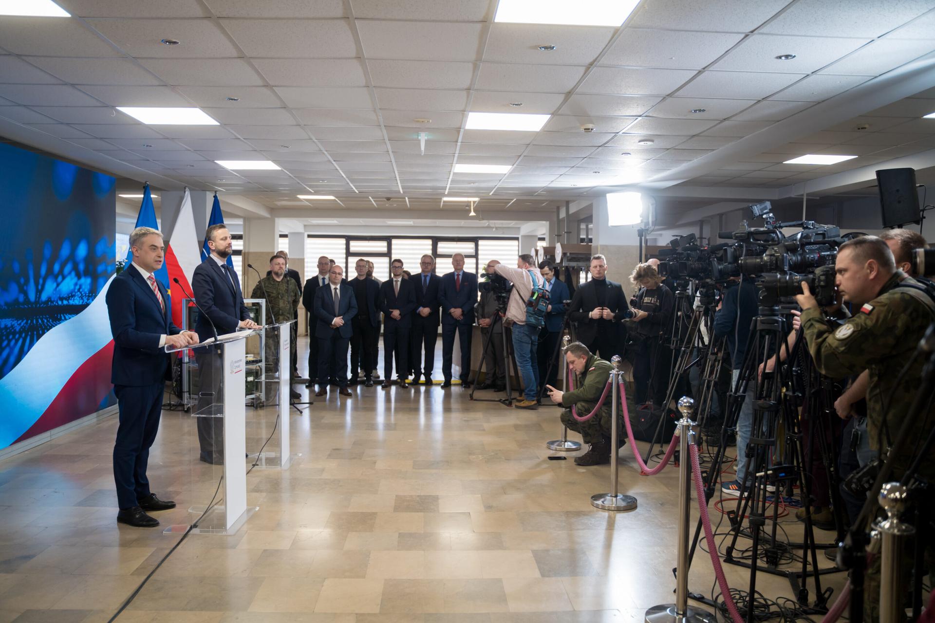 Wicepremier i Minister Cyfryzacji Krzysztof Gawkowski oraz Wicepremier i Minister Obrony Narodowej Władysław Kosiniak-Kamysz przemawiają podczas konferencji prasowej. Na sali obecni są dziennikarze, przedstawiciele mediów z kamerami oraz goście w tym osoby w mundurach wojskowych i garniturach. W tle widoczne są flagi Polski. Pomieszczenie jest jasne, przestronne, z ustawionymi mikrofonami i pulpitami dla przemawiających.