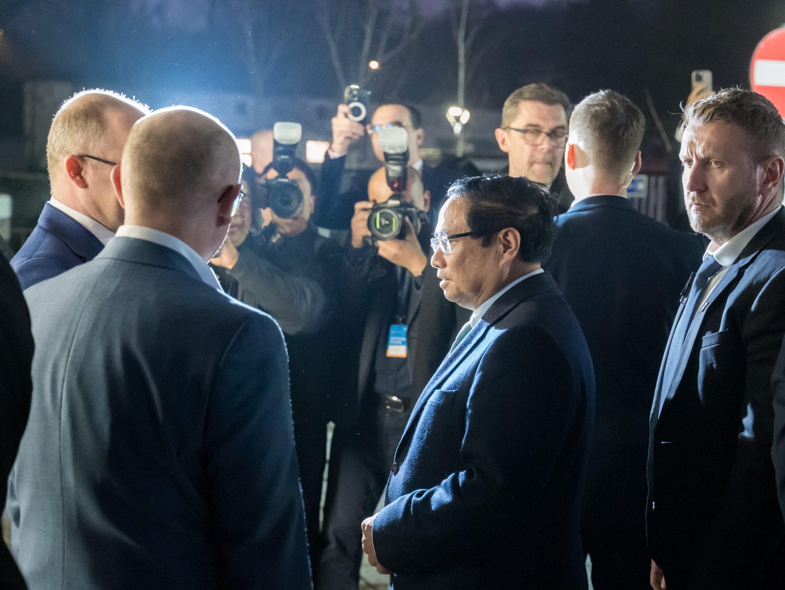 Premier Wietnamu Phạm Minh Chính stoi, otoczony przez ludzi witających go w świetle aparatów fotograficznych. Po lewej stronie znajduje się Paweł Olszewski, sekretarz stanu w Ministerstwie Cyfryzacji, a obok niego dyrektor NASK Radosław Nielek. W tle widać fotografów i dziennikarzy robiących zdjęcia oraz nocną scenerię.