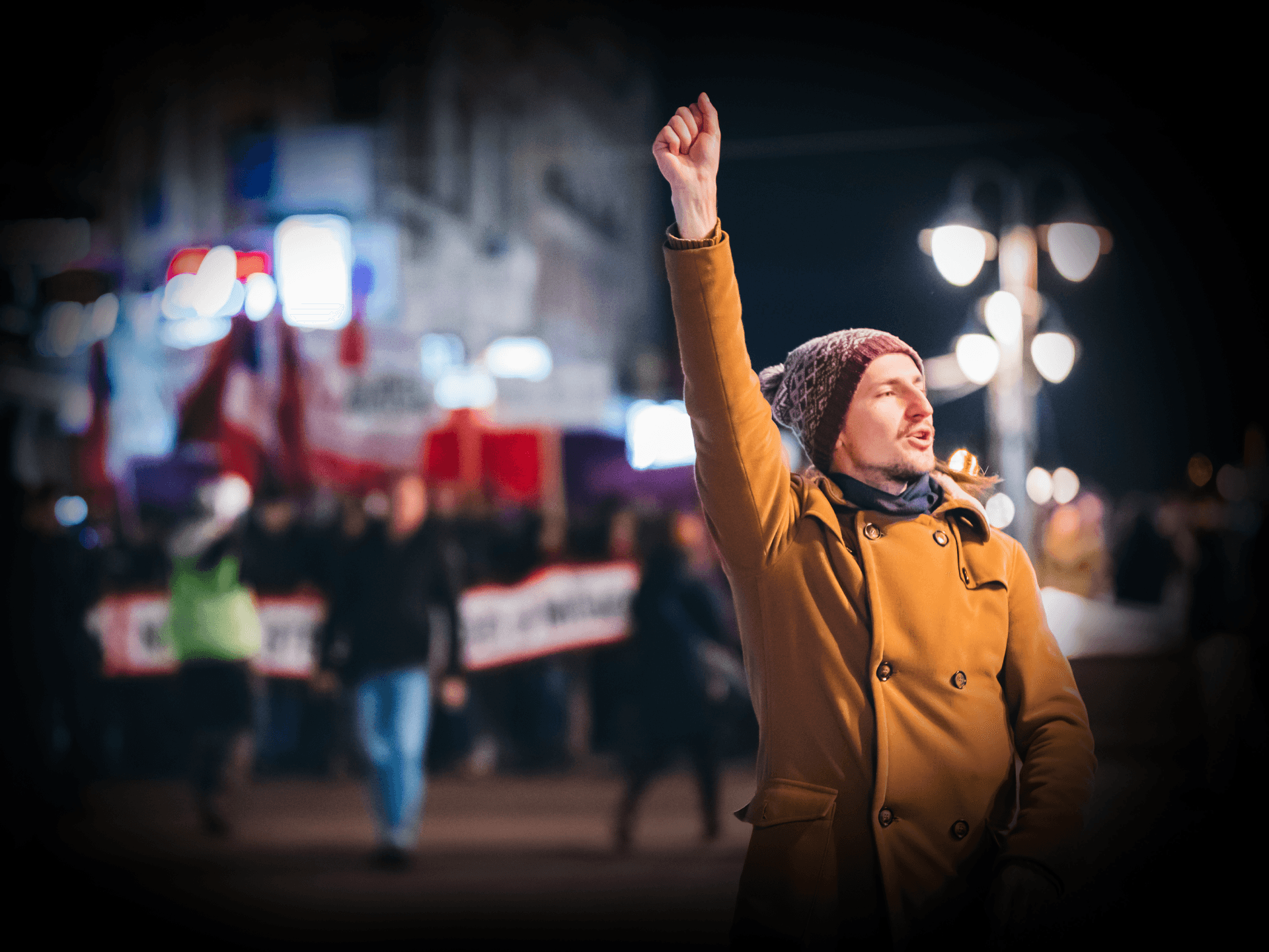 Mężczyzna wyciąga do góry rękę z dłonią zaciśniętą w pięść. W tle widać pochód ludzi z hasłami.