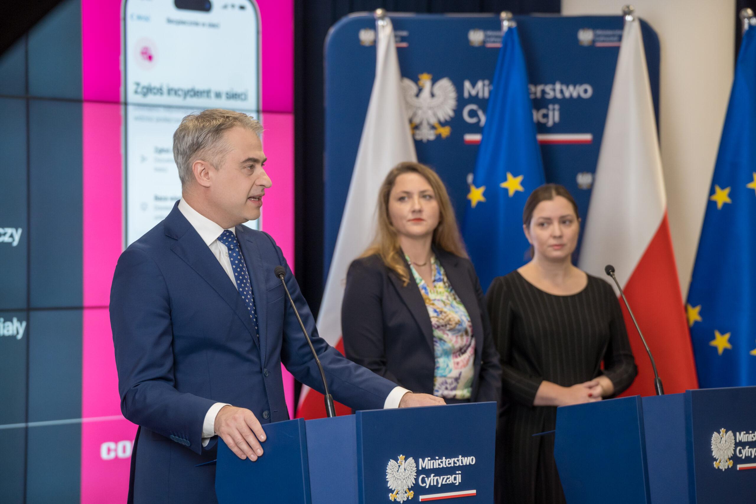 Mężczyzna (K. Gawkowski) stoi przy mównicy z napisem Ministerstwo cyfryzacji, za nim przy drugiej mównicy dwie kobiety słuchające tego co mówi.