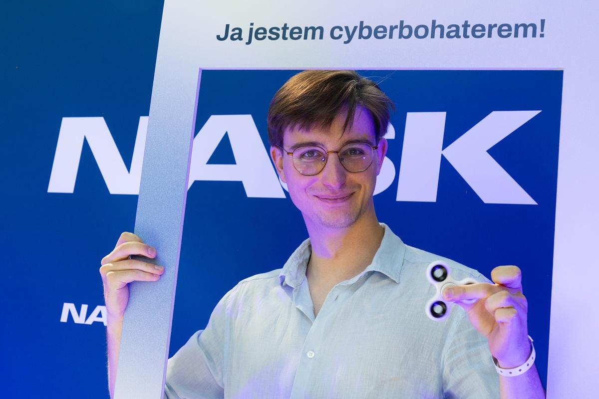 Na zdjęciu Karol Bojke, specjalista ds. incydentów i komunikacji w obszarze cyberbezpieczeństwa w CERT Polska. Uśmiechnięty, trzyma w rękach ramkę z napisem „Ja jestem cyberbohaterem!” oraz biały fidget spinner. W tle widoczne logo NASK na niebieskim tle.