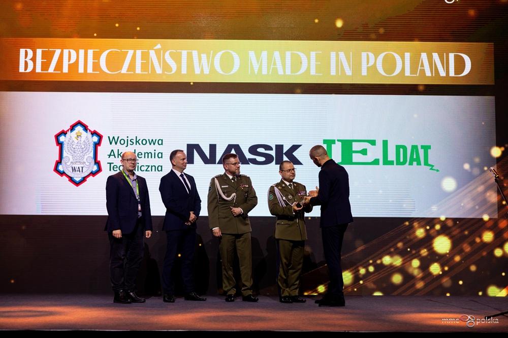 Grupa pięciu mężczyzn, w tym dwóch w mundurach wojskowych i trzech w garniturach, stoi na scenie podczas ceremonii wręczenia nagród. Jeden z wojskowych odbiera nagrodę od mężczyzny w granatowym garniturze. W tle widoczny jest ekran z napisem "BEZPIECZEŃSTWO MADE IN POLAND" oraz logotypami Wojskowej Akademii Technicznej (WAT), NASK i TELDAT. Scena jest oświetlona światłem reflektorów, a tło zawiera złote efekty świetlne.
