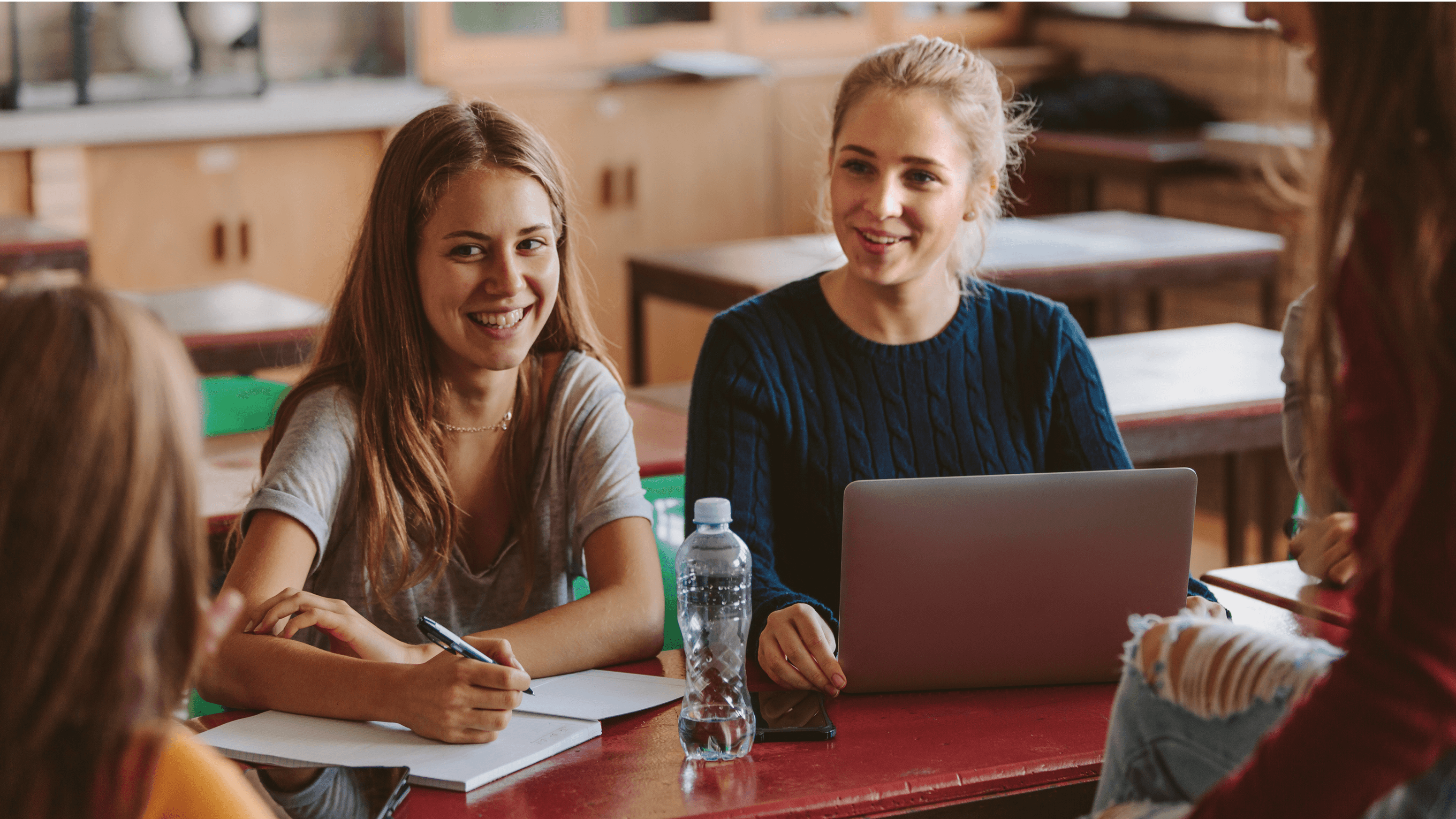 Dwie dziewczyny siedzące przy biurku w klasie, jedna pisze w zeszycie, druga korzysta z komputera.