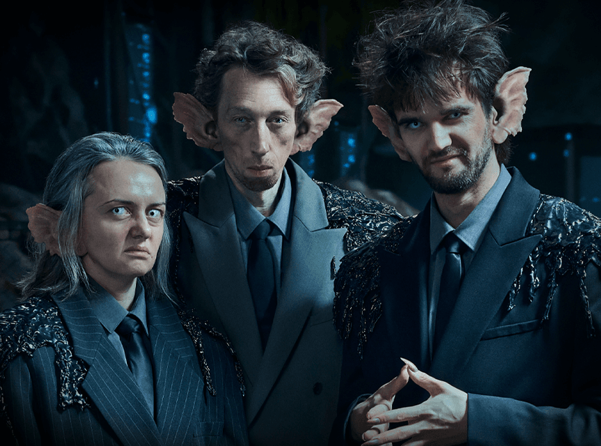 Trzy postaci, jedna kobieta i dwóch mężczyzn, z wykrzywionymi twarzami, nastroszonymi włosami i wielkimi uszami trolli. Mają miny jakby coś knuły, jeden z mężczyzn zaciera ręce z długimi szponami.