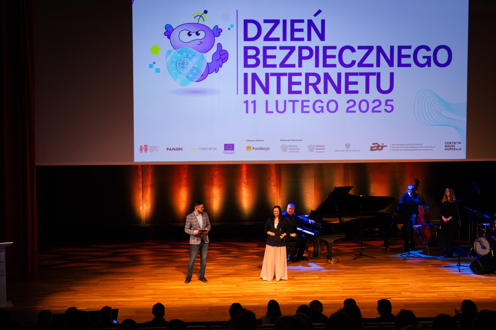 Fotografia przedstawia wydarzenie na scenie podczas obchodów Dnia Bezpiecznego Internetu 11 lutego 2025 roku. Na dużym ekranie w tle widnieje kolorowy baner z napisem 'Dzień Bezpiecznego Internetu' oraz grafiką fioletowej maskotki trzymającej tarczę z symbolem internetu. Na scenie znajduje się dwoje prowadzących. W głębi sceny stoi fortepian, za którym siedzi pianista, a po prawej stronie występuje kilku muzyków, w tym kontrabasista i wokalistka. Cała scena jest podświetlona ciepłym, pomarańczowym światłem, a widownia widoczna jest w dolnej części obrazu.