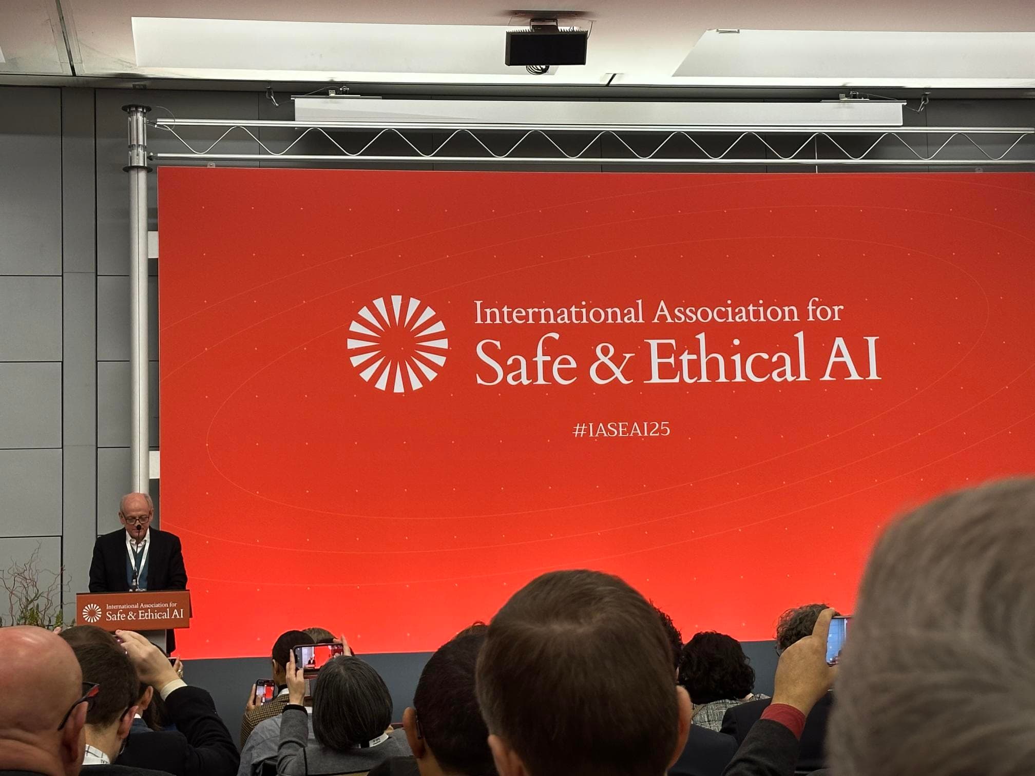 Na zdjęciu Joseph E. Stiglitz, laureat Nagrody Nobla w dziedzinie ekonomii, siedzi na scenie podczas konferencji International Association for Safe & Ethical AI (#IASEAI25). W tle widoczna jest czerwona ścianka z logo wydarzenia. Stiglitz trzyma mikrofon i wygląda na skoncentrowanego, a publiczność uważnie słucha jego wypowiedzi. W kadrze widać także mikrofon na statywie i osobę nagrywającą wystąpienie telefonem."