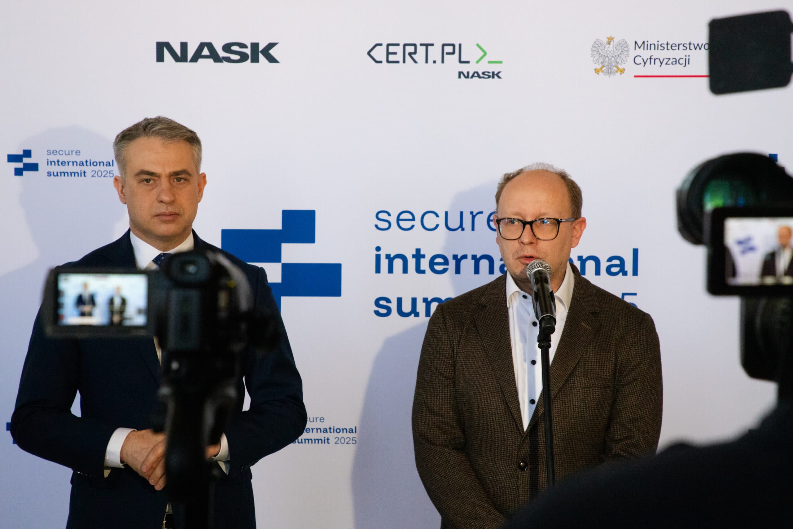 Dwaj mężczyźni stojący na tle ścianki konferencyjnej z napisem „Secure International Summit 2025”. Po lewej stronie stoi Krzysztof Gawkowski, wicepremier i minister cyfryzacji, ubrany w ciemny garnitur. Po prawej stronie przy mikrofonie stoi Radosław Nielek, dyrektor NASK, w ciemnej marynarce i jasnej koszuli. Przed nimi ustawione są kamery, trwa konferencja prasowa.