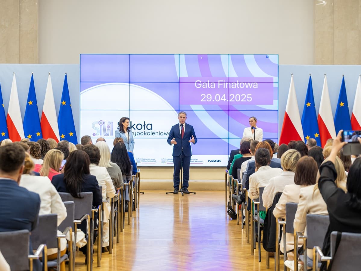 Na zdjęciu widać uroczystość finałową programu "Szkoła Pokoleń", która odbyła się 29 kwietnia 2025 roku. Przemawiają trzy osoby stojące na scenie przed dużym ekranem z napisem "Gala Finałowa 29.04.2025". Od lewej strony widoczna jest Marzena Okła-Drewnowicz, ministra ds. polityki senioralnej, w środku przemawia wicepremier i minister cyfryzacji Krzysztof Gawkowski, a po prawej Barbara Nowacka, ministra edukacji. Publiczność, złożona z uczestników wydarzenia, siedzi tyłem do aparatu. Tło zdobią flagi Polski i Unii Europejskiej.
