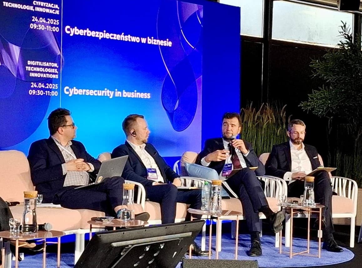 Maciej Siciarek, dyrektor CSIRT, wypowiada się podczas panelu dyskusyjnego na temat „Cyberbezpieczeństwo w biznesie” w ramach wydarzenia „Cyfryzacja, Technologie, Innowacje” 24 kwietnia 2025 roku. Obok niego siedzi trzech innych uczestników panelu, wszyscy w garniturach, z mikrofonami i laptopami. W tle widać niebieski ekran z nazwą wydarzenia w języku polskim i angielskim.
