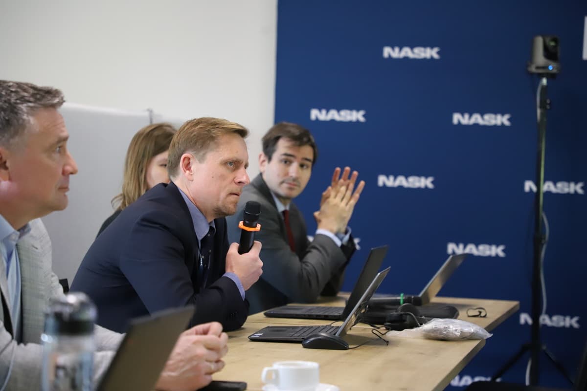 Paweł Kostkiewicz, dyrektor ds. certyfikacji w NASK, zabiera głos podczas spotkania, trzymając mikrofon. W tle widoczny jest baner z logo NASK, a przy stole siedzą również inni uczestnicy z laptopami.