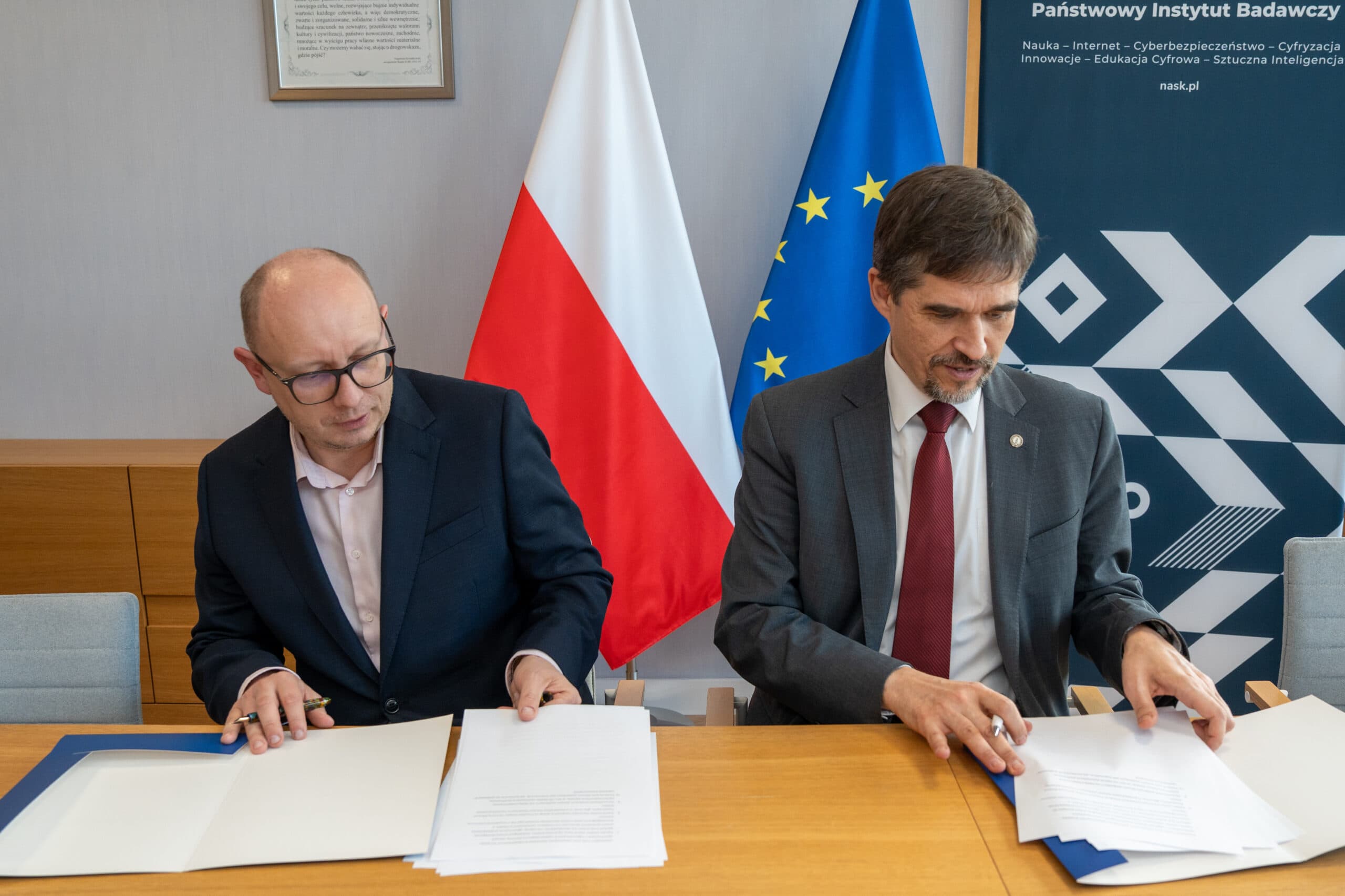 Dwóch mężczyzn w garniturach siedzi przy stole i przegląda dokumenty, za nimi stoją flagi Polski i Unii Europejskiej.
