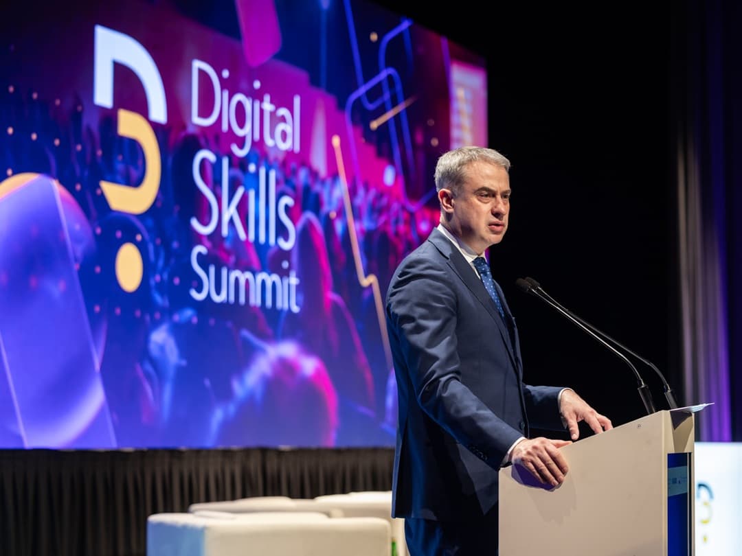 Krzysztof Gawkowski stoi przy mównicy i przemawia podczas konferencji. W tle znajduje się duży, kolorowy ekran z napisem „Digital Skills Summit” i graficznymi, abstrakcyjnymi efektami w odcieniach fioletu, różu i niebieskiego. Obok mównicy ustawione są mikrofony, a przed sceną widać jasne, tapicerowane siedziska.