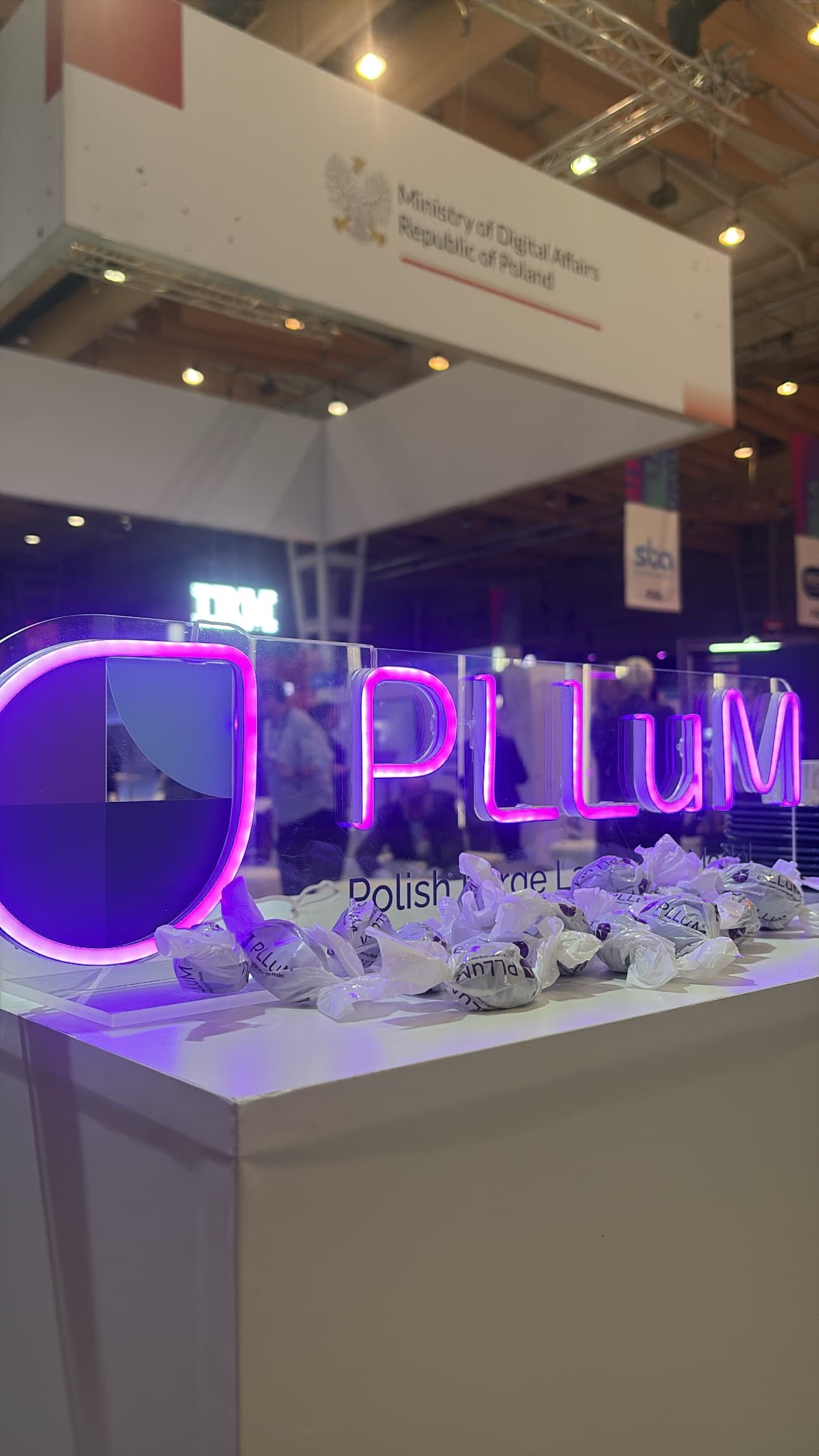 Zbliżenie na podświetlone logo PLLuM – Polish Large Language Model – na stoisku Polski podczas międzynarodowych targów technologicznych. W tle widoczne oznaczenia Ministerstwa Cyfryzacji.