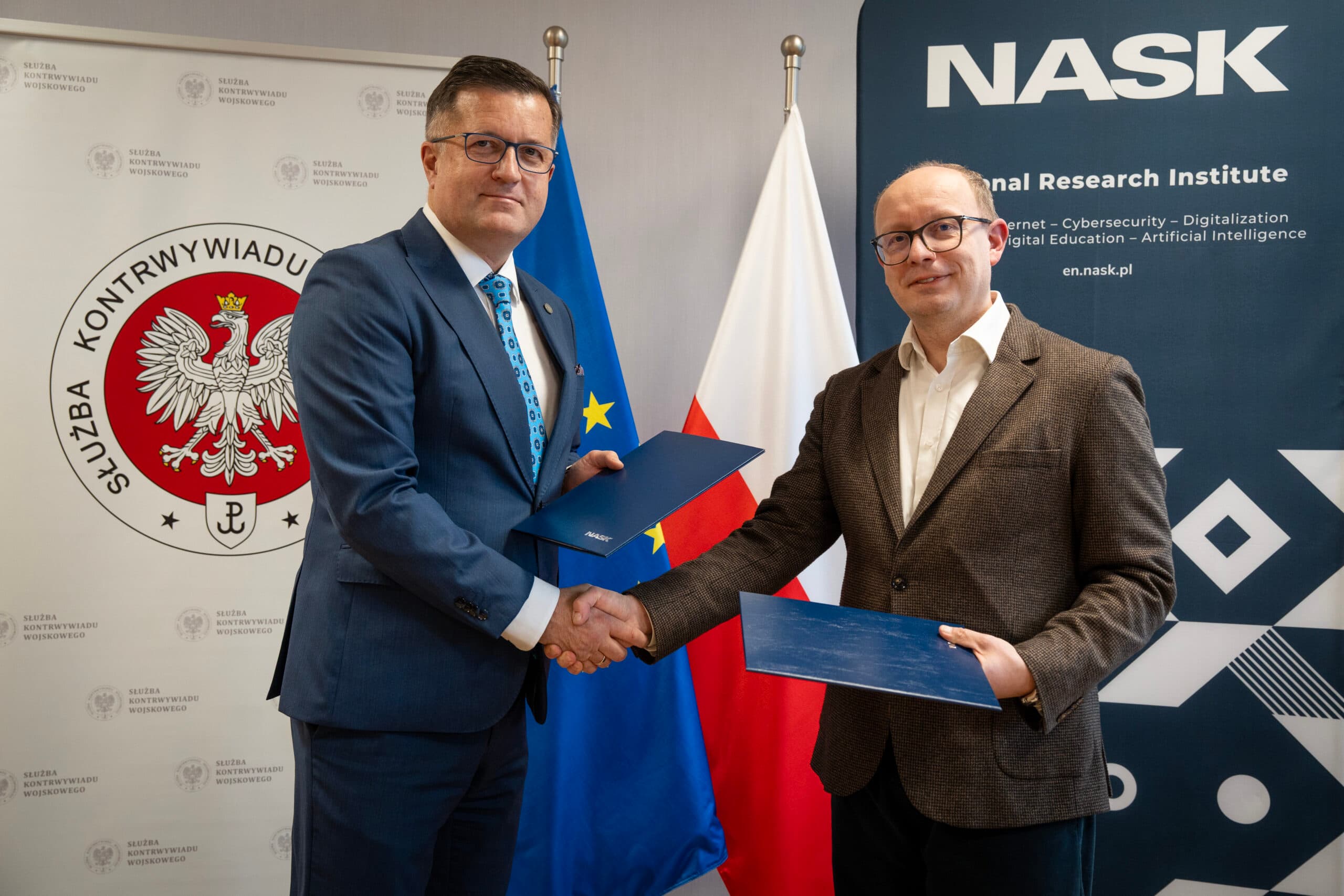 Dwaj mężczyźni ściskają sobie dłonie po podpisaniu umowy o współpracy między NASK a Służbą Kontrwywiadu Wojskowego. W tle widoczne są flagi Polski i Unii Europejskiej oraz logotypy obu instytucji.