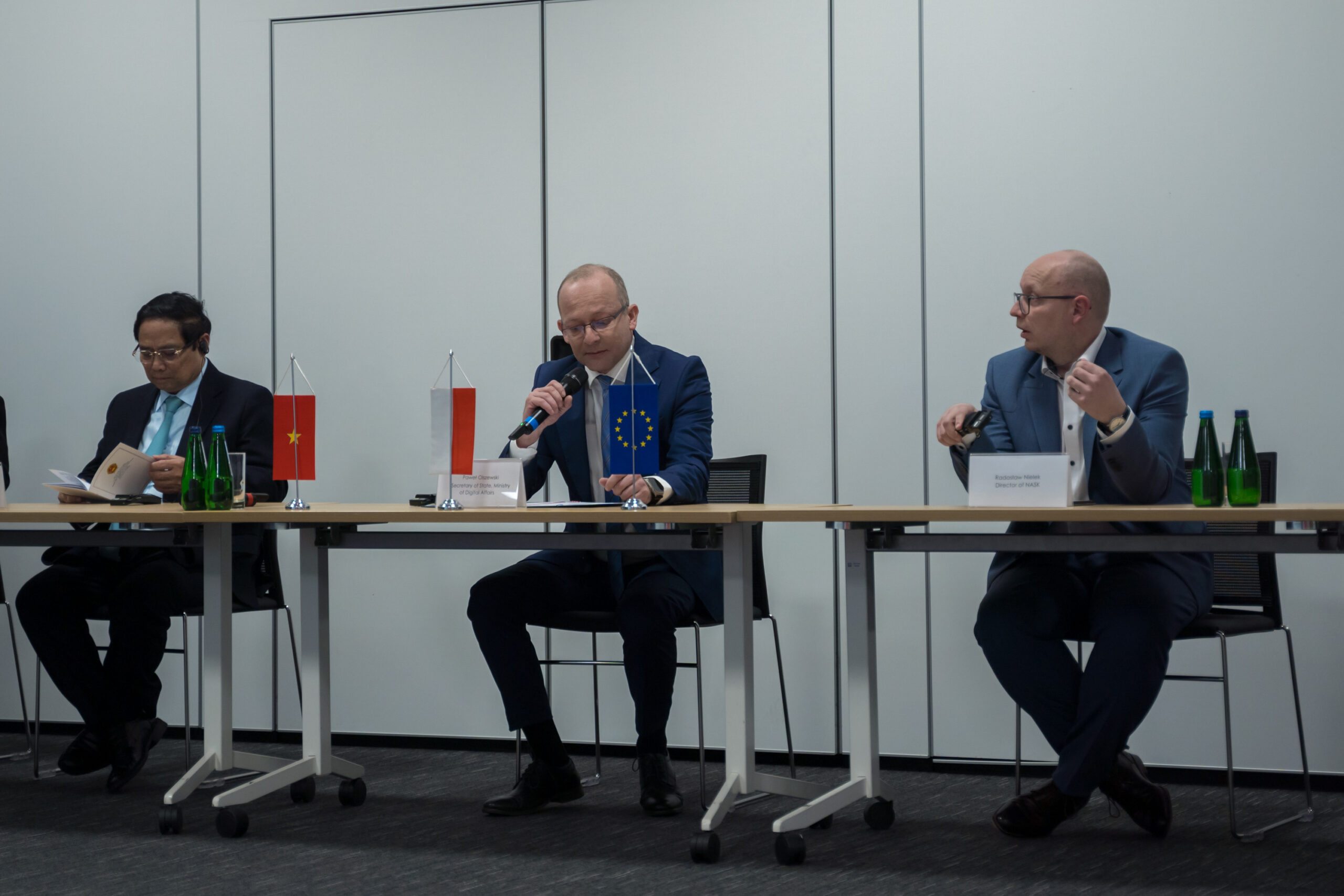 Na zdjęciu przy stole konferencyjnym siedzą trzy osoby: po lewej stronie Premier Wietnamu Phạm Minh Chính przegląda dokumenty, w środku Paweł Olszewski, sekretarz stanu w Ministerstwie Cyfryzacji, przemawia do mikrofonu, a po prawej stronie dyrektor NASK Radosław Nielek. Na stole widoczne są małe flagi Wietnamu, Polski i Unii Europejskiej.