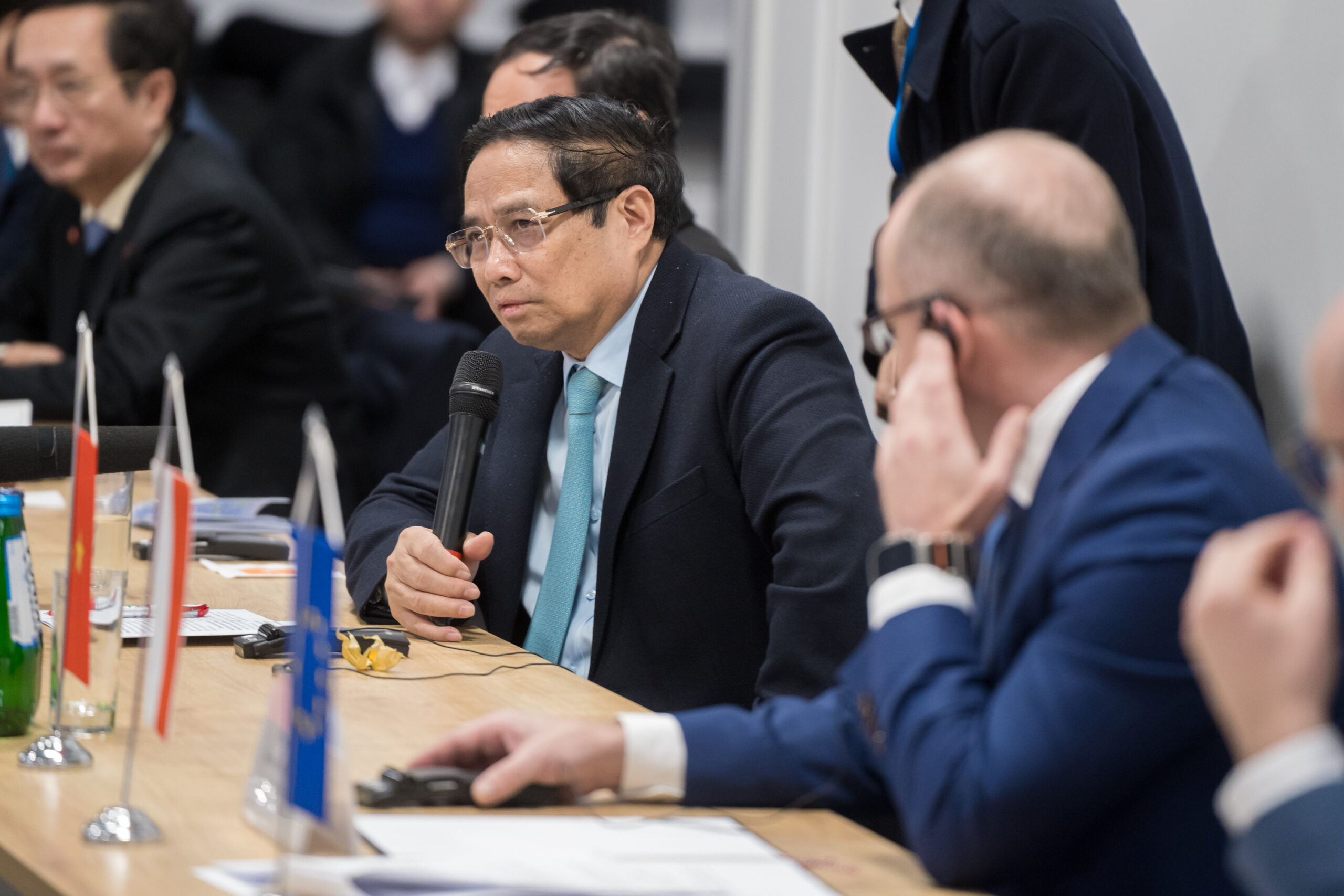Premier Wietnamu Phạm Minh Chính siedzi przy stole konferencyjnym, trzymając mikrofon i wygłaszając przemówienie lub odpowiadając na pytanie. W tle widać innych uczestników spotkania, a na stole znajdują się małe flagi symbolizujące Wietnam, Polskę i Unię Europejską.