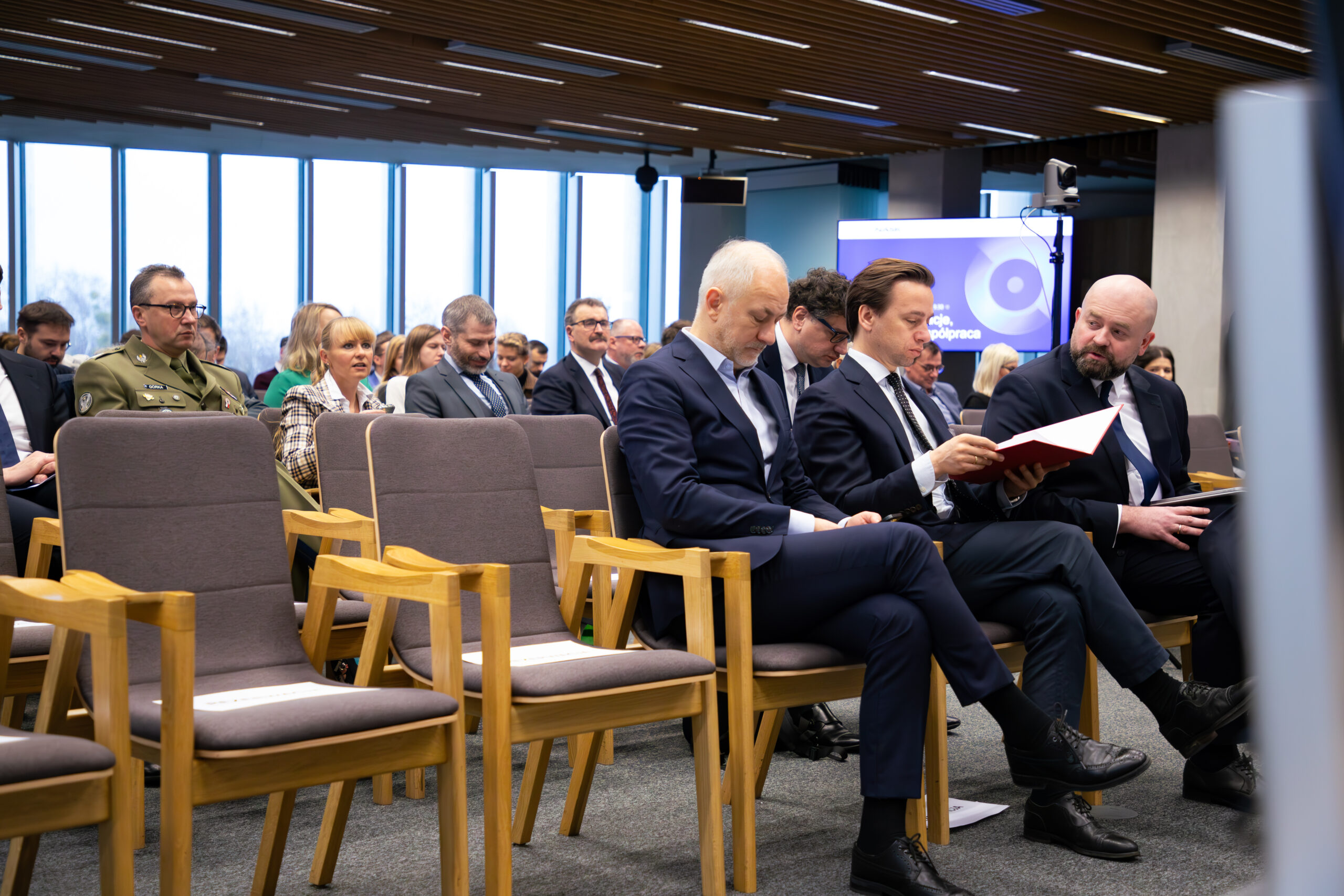 Na zdjęciu uwieczniono konferencję odbywającą się w Sejmie RP. W pierwszym rzędzie siedzą Bartłomiej Pejo, Krzysztof Bosak oraz Grzegorz Napieralski. Ubrani są w formalne stroje, trzymają notatki. W tle widać innych uczestników konferencji, a także ekran z fragmentem napisu wskazującego temat spotkania. Sala ma nowoczesny wystrój z dużymi oknami zapewniającymi dostęp do naturalnego światła.