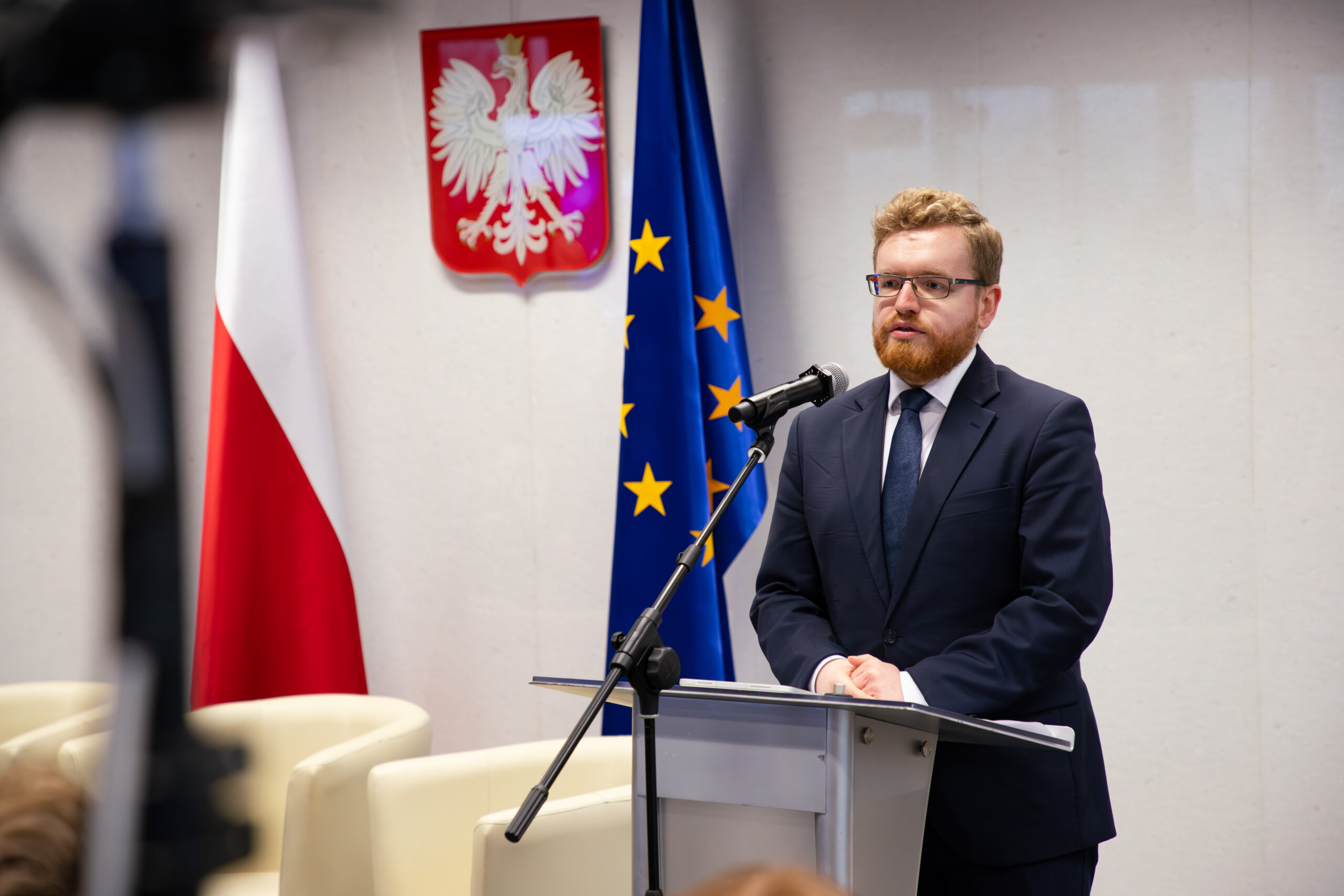 Na zdjęciu widoczny jest Dariusz Standerski podczas przemówienia na konferencji w Sejmie RP. Stoi przy mównicy, ubrany w elegancki garnitur, i przemawia do zgromadzonych uczestników. W tle znajdują się flaga Polski, flaga Unii Europejskiej oraz godło Polski, co podkreśla oficjalny charakter wydarzenia. Widać mikrofon ustawiony na mównicy, co wskazuje na formalną formę wystąpienia.