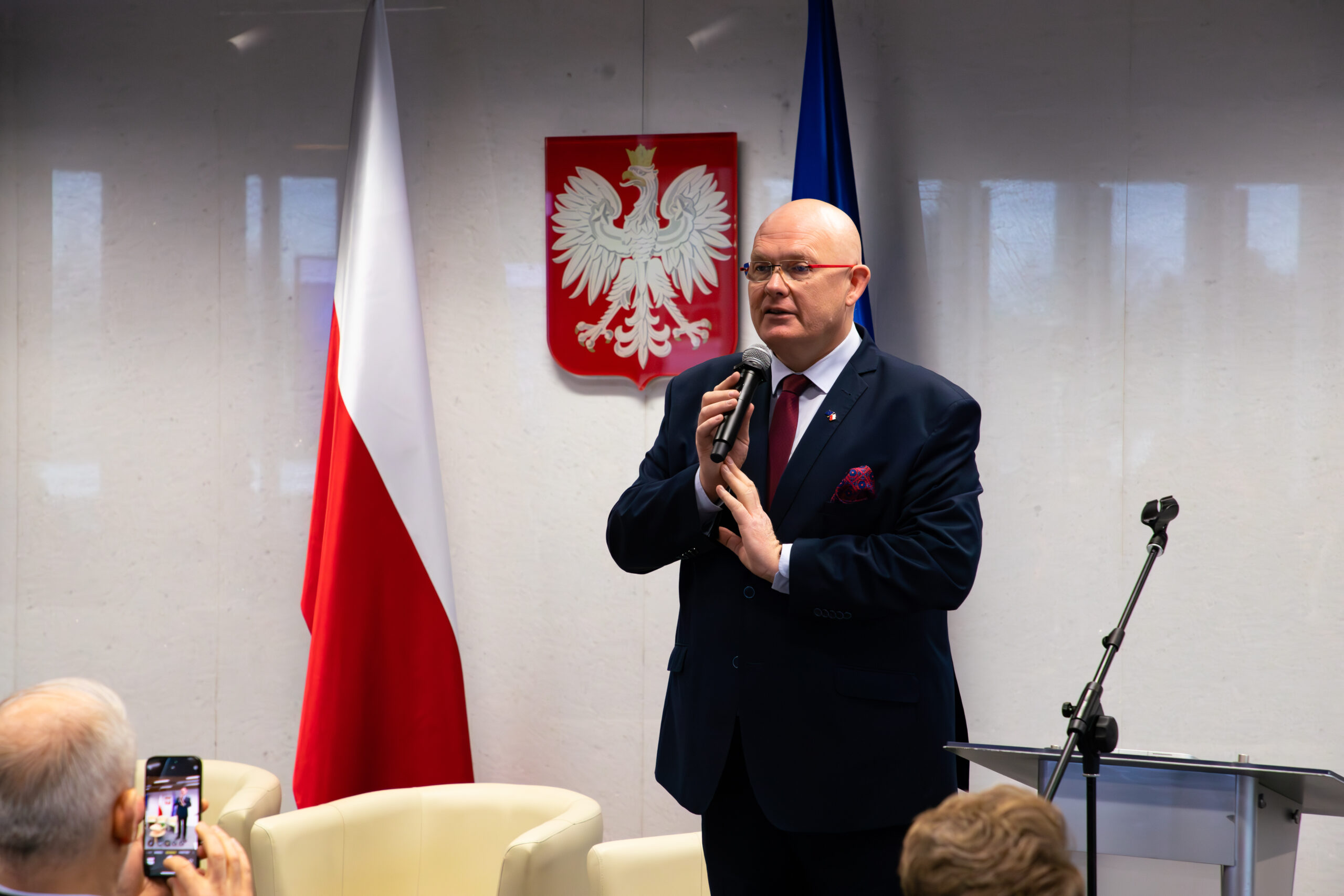 Na zdjęciu uwieczniono Michała Gramatykę podczas wystąpienia na konferencji w Sejmie RP. Mężczyzna ubrany w elegancki garnitur, trzyma mikrofon, przemawiając do uczestników. W tle widoczne są flagi Polski i Unii Europejskiej oraz godło Polski na ścianie, co podkreśla oficjalny charakter wydarzenia. Na pierwszym planie widać fragment publiczności, w tym osobę robiącą zdjęcie telefonem.