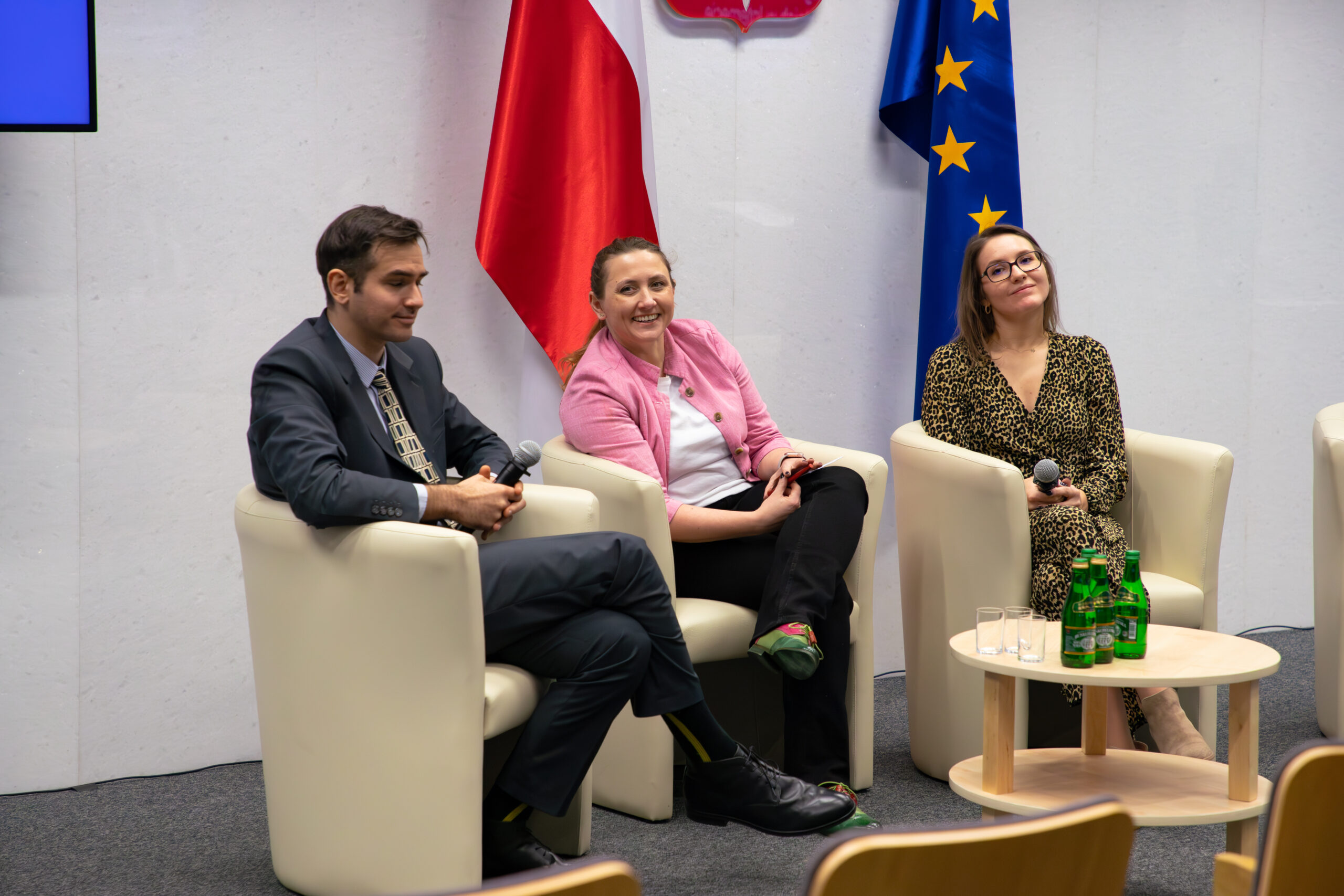 Na zdjęciu widoczna jest scena konferencji odbywającej się w Sejmie RP. Trzy osoby siedzą na fotelach: Martyna Różycka, Ewelina Bartuzi-Trokielewicz oraz Filip Konopczyński. W tle widoczne są flagi Polski i Unii Europejskiej, a uczestnicy biorą udział w dyskusji panelowej.