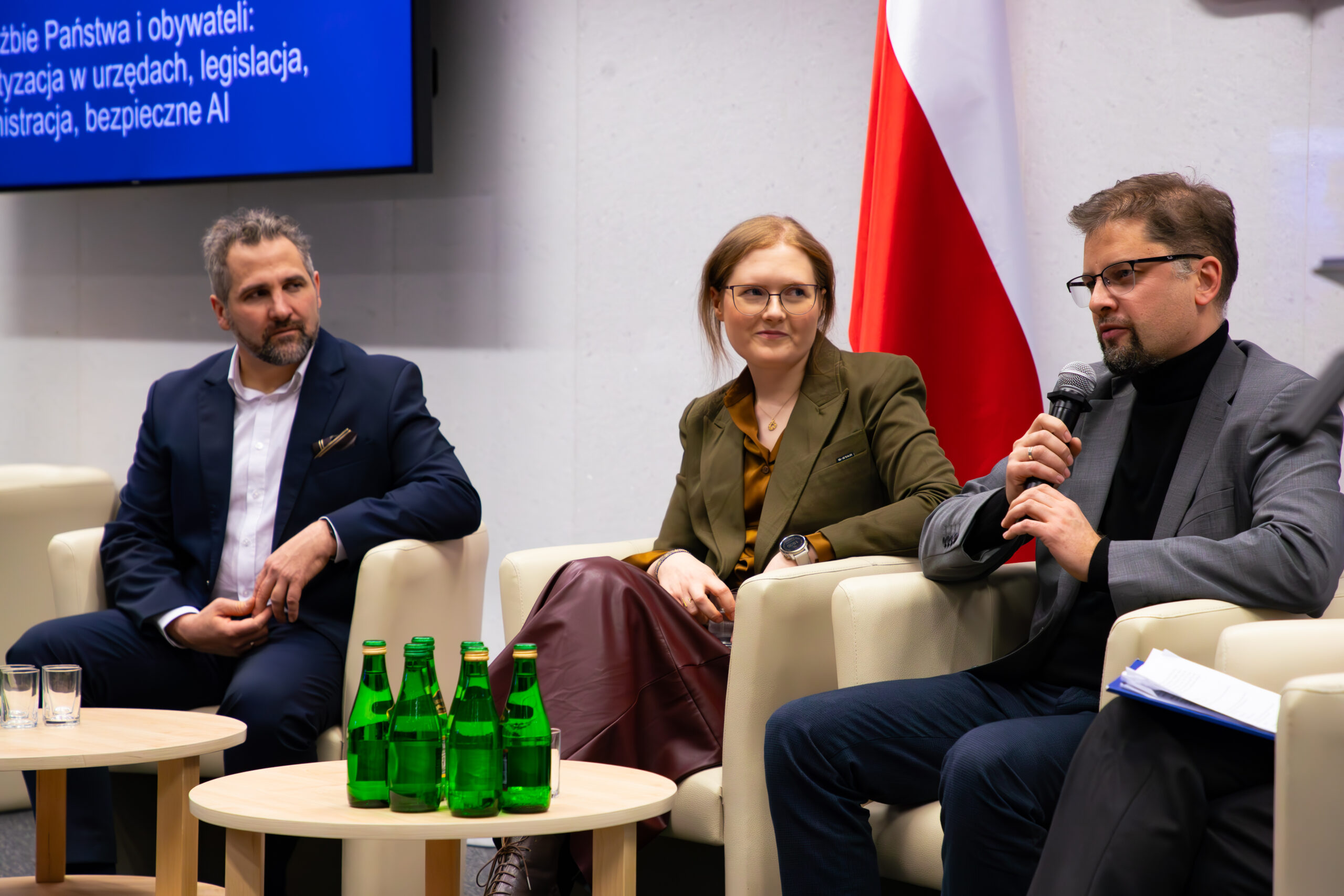 Na zdjęciu uwieczniono panel dyskusyjny podczas konferencji w Sejmie RP. Uczestnikami są Szymon Łukasik, Agnieszka Karlińska i Grzegorz Żmijewski, siedzący na białych fotelach. Szymon Łukasik trzyma mikrofon i wygłasza swoją wypowiedź. W tle widoczna jest flaga Polski oraz ekran z fragmentem napisu dotyczącym tematu dyskusji: „...bezpieczne AI”.